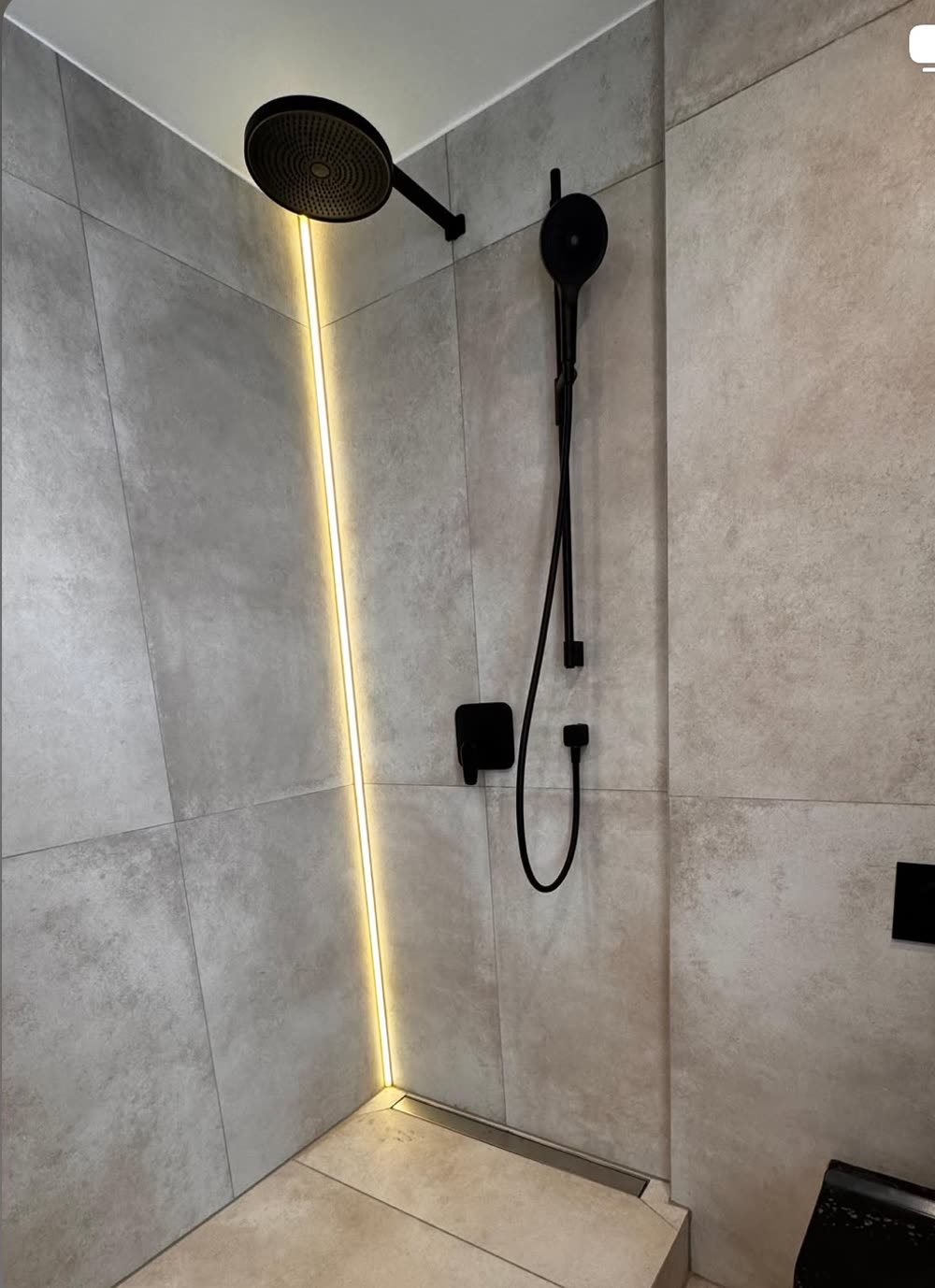 Fertig sanierte bodengleiche Dusche mit großformatigen Fliesen in Betonoptik, schwarzen Armaturen und einem LED-Lichtband in der Ecke.