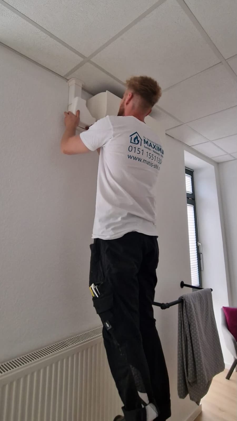 Maxim Matis montiert einen Lüftungsauslass an der Decke, Firmenshirt MAXIMal sichtbar